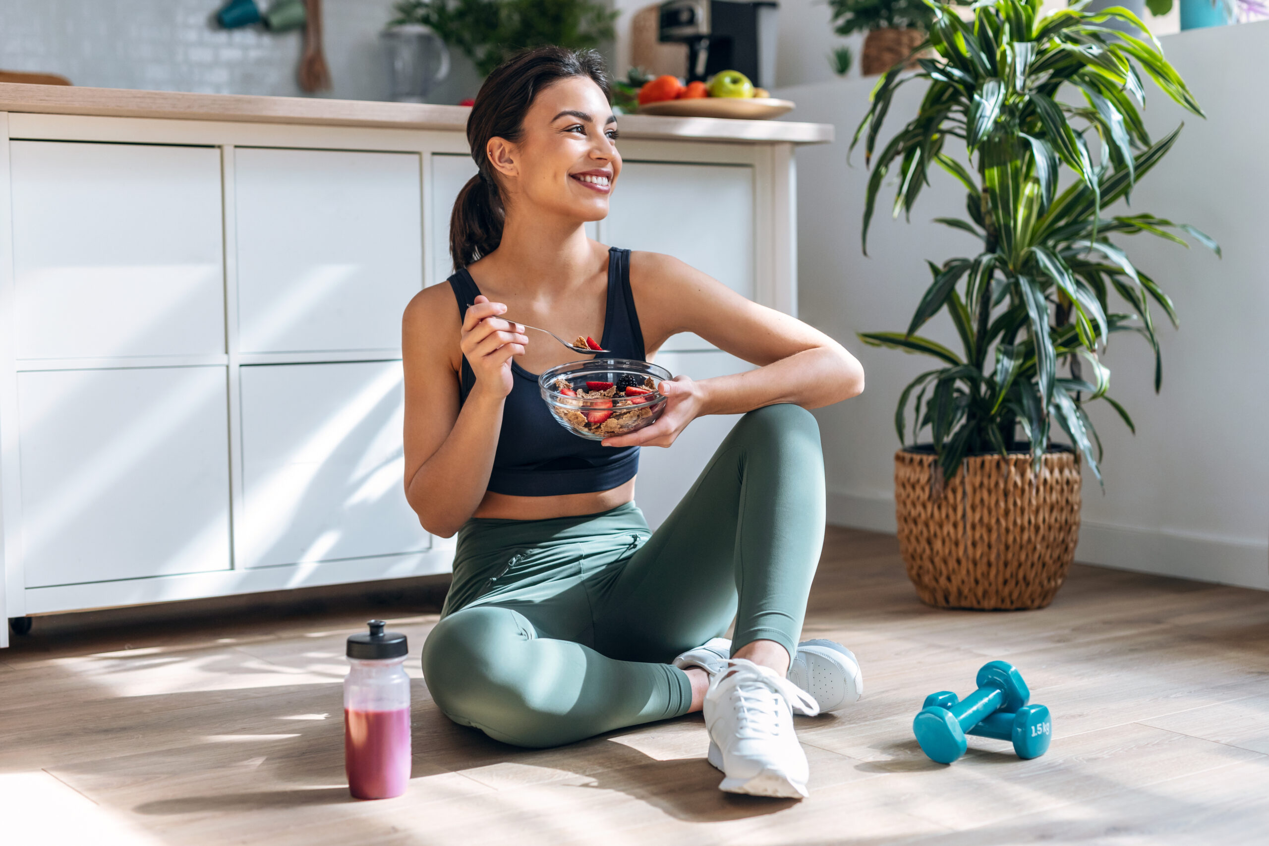 athletic woman eating a healthy bowl of muesli with fruit sitting on floor in the kitchen at home